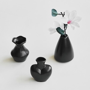May include: Three small matte black ceramic vases of varying shapes. One vase is heart-shaped, one has a ruffled rim, and the tallest vase holds a white magnolia flower with green leaves. The vases are arranged on a plain, light-colored surface.