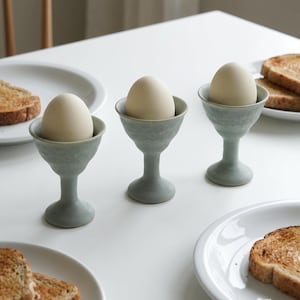 Könnte beinhalten: Drei hellblaue Keramik-Eierbecher, die jeweils ein weißes Ei enthalten, sind auf einer weißen Oberfläche angeordnet. Toastscheiben sind auf weißen Tellern zu sehen, was auf ein Frühstück hindeutet.