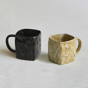 May include: Two square-shaped ceramic mugs. One is black, the other is beige with a marbled effect. Both mugs have a textured surface and a curved handle. The mugs are displayed on a white surface.
