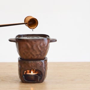 May include: A dark brown ceramic fondue pot with two side handles sits atop a matching base containing a lit tea light candle. A wooden ladle is positioned above the pot, with a single drop of liquid falling into the centre of the vessel.