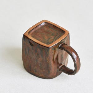 Może przedstawiać: Kwadratowy kubek ceramiczny z błyszczącym, nakrapianym szkliwem w odcieniach brązu i ciemnej miedzi. Kubek posiada grube, zakrzywione ucho z jednej strony oraz płaską, kwadratową podstawę. Powierzchnia ma nier&oacute;wną, teksturowaną strukturę.