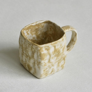 Może przedstawiać: Kwadratowy ceramiczny kubek z marmurkowym wzorem w odcieniach brązu i bieli. Kubek posiada zaokrąglone ucho po prawej stronie oraz teksturowaną, nier&oacute;wną powierzchnię, kt&oacute;ra nadaje mu wyrazisty, organiczny wygląd na całej zewnętrznej stronie.