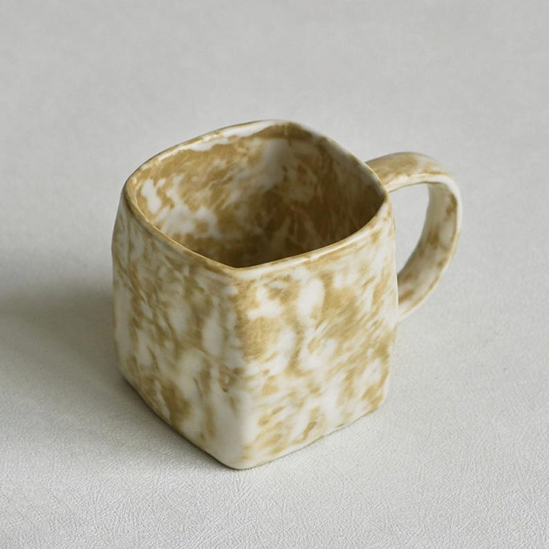 Może przedstawiać: Kwadratowy ceramiczny kubek z marmurkowym wzorem w odcieniach brązu i bieli. Kubek posiada zaokrąglone ucho po prawej stronie oraz teksturowaną, nier&oacute;wną powierzchnię, kt&oacute;ra nadaje mu wyrazisty, organiczny wygląd na całej zewnętrznej stronie.