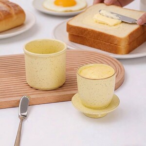 Può includere: Due tazze in ceramica giallo pallido con finitura maculata. Una tazza è vuota e cilindrica, mentre l'altra è riempita di burro e poggia su un piccolo piattino circolare abbinato. Un coltello da burro in metallo argentato è posizionato a sinistra.