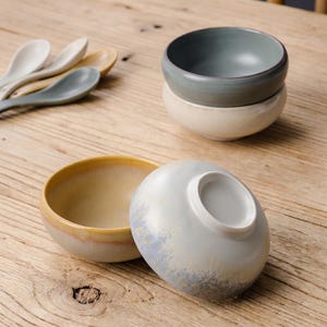 May include: A collection of ceramic bowls and spoons in various colors. The bowls are in shades of gray, beige, and yellow. The spoons are in white, yellow, and gray. The bowls are stacked and arranged on a wooden surface.