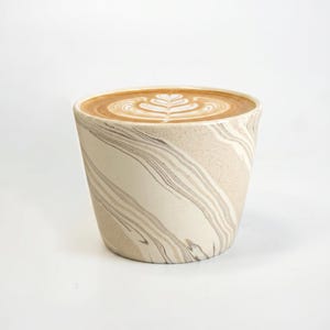 May include: A ceramic cup with a speckled cream and brown marbled swirl pattern filled with a coffee beverage featuring a white rosetta latte art design on the surface. The cup has a tapered cylindrical shape and a smooth, matte-like finish.