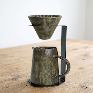 May include: A ceramic coffee dripper with a textured olive green finish and vertical grooves, suspended above a matching ceramic pitcher with a handle, both supported by a black metal stand with a flat base and a vertical arm.