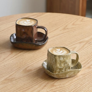 May include: Two square ceramic cups with matching saucers, each containing a latte with heart-shaped foam art. One cup is a dark, glossy brown, while the other is a textured, mottled light olive green. Both feature irregular, hand-formed edges.