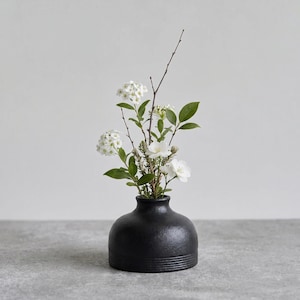 Puede incluir: Un jarrón de cerámica negro mate con cuerpo redondeado y base texturizada con estrías. El jarrón contiene un pequeño arreglo de flores blancas, hojas verdes y ramas delgadas y desnudas de color marrón, contrastando con el recipiente oscuro.