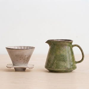 May include: A ceramic coffee dripper with a light gray, speckled finish and a fluted interior, paired with a matching green, textured ceramic pitcher featuring a wide base, a curved handle, and a distinct pouring spout for liquid service.