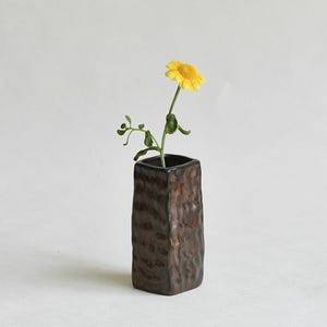 May include: A rectangular ceramic vase with a dark brown, textured, and dimpled surface. A single yellow flower with green leaves and a thin stem is placed inside the vase, extending upwards from the opening at the top of the vessel.