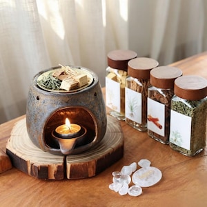 May include: A dark grey ceramic oil burner with a lit tea light candle inside, topped with a brass dish containing dried herbs and wood pieces. Beside it are four glass jars with wooden lids filled with dried botanicals and clear quartz stones.