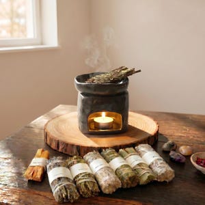 May include: A black ceramic oil burner with a gold interior holds a lit tea light candle and a bundle of dried herbs. In front, seven tied bundles of dried herbs with white paper labels are arranged on a round wooden slice with natural bark edges.