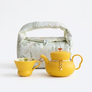 May include: A bright yellow teapot with a pearl-accented band and a leaf-shaped charm, paired with a matching yellow teacup. Behind them sits a padded fabric bag featuring a light green and white botanical print with a single button closure.