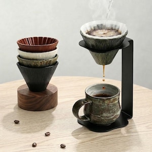 Puede incluir: Un juego de cuatro goteros de café de cerámica estriada en marrón, blanco, verde y negro, apilados sobre una base de madera. Un soporte de metal negro sostiene un gotero verde con filtro de papel, vertiendo café en una taza de cerámica verde.