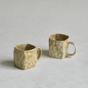 Może przedstawiać: Dwa kwadratowe kubki ceramiczne z marmurkowym wzorem w odcieniach brązu i bieli. Każdy kubek ma zaokrąglone ucho z boku oraz teksturowaną, nier&oacute;wną powierzchnię. Kubki są ustawione obok siebie, prezentując sp&oacute;jny wygląd i formę wykonania.