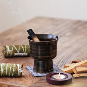 Pode incluir: Um queimador de incenso de cerâmica castanho-escuro em forma de cálice contendo um pedaço de madeira de palo santo fumegante com fumo visível. Dois molhos de sálvia verdes envolvidos em fio branco e vários paus de madeira clara estão próximos.