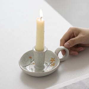 Puede incluir: Un candelabro de cerámica de color blanco roto con motas, un asa circular y un soporte central. La base presenta motivos florales pintados a mano en amarillo y naranja. Una vela cónica de color crema está insertada en el soporte y encendida.