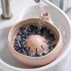 May include: A round, light pink ceramic berry colander bowl with a central drainage dome and a side handle, filled with fresh blueberries being rinsed under a stream of water. The bowl features a smooth, matte finish and a wide, open top design.