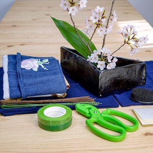 Puede incluir: Un jarrón de cerámica negro rectangular con ramas de cerezo, una toalla azul bordada, cinta floral verde, tijeras florales verdes, alambres florales finos marrones y verdes y un alfiletero floral negro sobre un paño azul.
