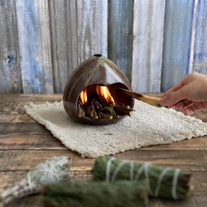 May include: A dark brown, teardrop-shaped ceramic incense burner with a wide opening, containing burning wood sticks. The burner sits on a textured, off-white fabric square, with two additional bundles of dried herbs placed in the foreground.