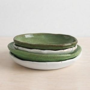 May include: A stack of four small ceramic plates with irregular, hand-formed edges. The plates alternate in color between a speckled moss green and a solid off-white glaze, featuring a smooth, glossy finish and a shallow, rounded basin shape.