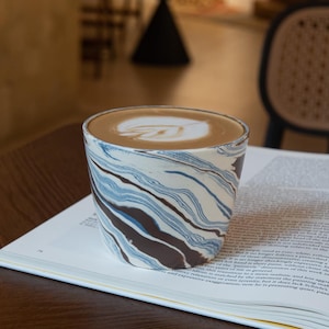 May include: A ceramic cup with a marbled pattern in shades of blue, white, and dark brown, filled with a latte featuring a white leaf-shaped foam design. The cup has a wide, rimless opening and a tapered base, showcasing a smooth, glossy finish.