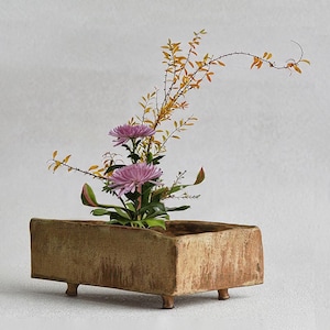 May include: A rectangular, tan-colored ceramic planter with a textured, rustic finish and four small feet. The planter holds a floral arrangement featuring two large, light purple chrysanthemum flowers and thin, arching branches with yellow leaves.