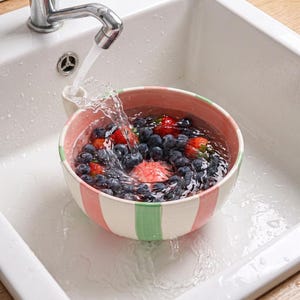 Puede incluir: Un cuenco de cerámica con rayas verticales rosas y verdes lleno de arándanos y fresas frescas siendo enjuagado bajo un chorro de agua de un grifo plateado. El cuenco está colocado dentro de un fregadero blanco con agua salpicando.