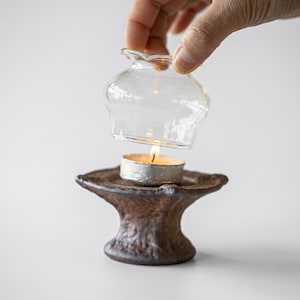 以下が含まれることがあります： 暗い茶色の質感のあるセラミック製キャンドルホルダー。幅広で広がったベースと上部が特徴です。中央には、火が灯った小さな銀色のティーライトキャンドルが置かれています。透明なベル型のガラスカバーが上にかざされています。