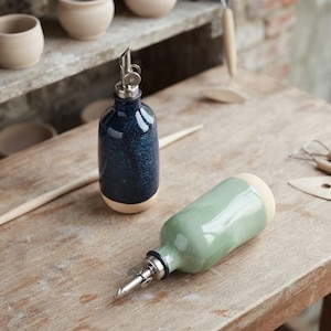May include: Two ceramic oil dispensers with stainless steel pour spouts. One bottle features a dark blue speckled glaze, while the other is finished in a light sage green glaze. Both bottles have a smooth, unglazed base and a rounded body shape.