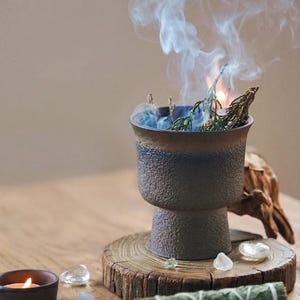Pode incluir: Um recipiente de cerâmica texturizada em forma de cálice com tons escuros e borda alargada, contendo ervas secas acesas que emitem uma coluna de fumaça branca. O recipiente apresenta um esmalte azul e marrom e repousa sobre uma base de madeira.
