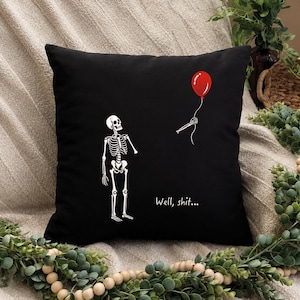 May include: A black square throw pillow featuring a white graphic of a skeleton looking at a detached skeletal hand holding a red balloon. The text "Well, shit..." is printed in white below the hand. The design is centered on the front of the pillow.
