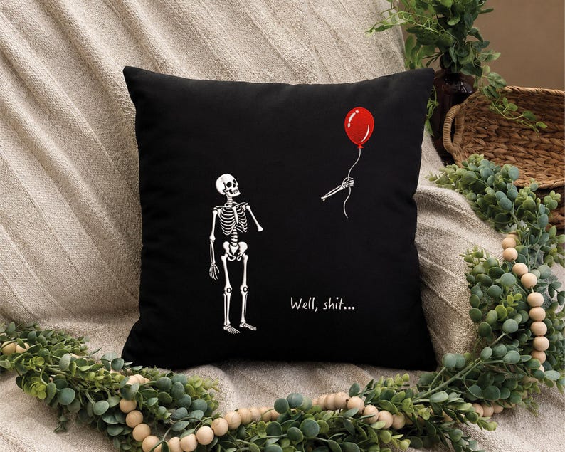 May include: A black square throw pillow featuring a white graphic of a skeleton looking at a detached skeletal hand holding a red balloon. The text "Well, shit..." is printed in white below the hand. The design is centered on the front of the pillow.