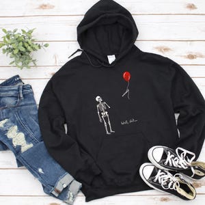 May include: A black hooded sweatshirt featuring a white graphic of a skeleton looking up at a red balloon floating away on a string. The text "Well, shit..." is printed in white below the skeleton. The hoodie has long sleeves and a front pocket.