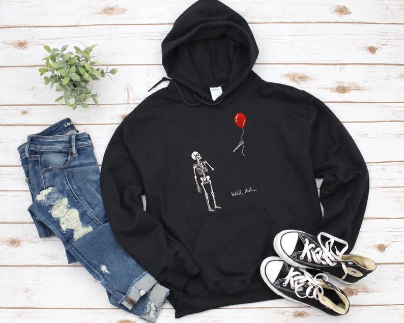 May include: A black hooded sweatshirt featuring a white graphic of a skeleton looking up at a red balloon floating away on a string. The text "Well, shit..." is printed in white below the skeleton. The hoodie has long sleeves and a front pocket.