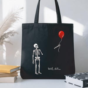 May include: A black tote bag featuring a white graphic of a skeleton looking up at a red balloon held by a disembodied skeletal hand. The text "Well, shit..." is printed in white below the skeleton. The bag has two black shoulder straps.