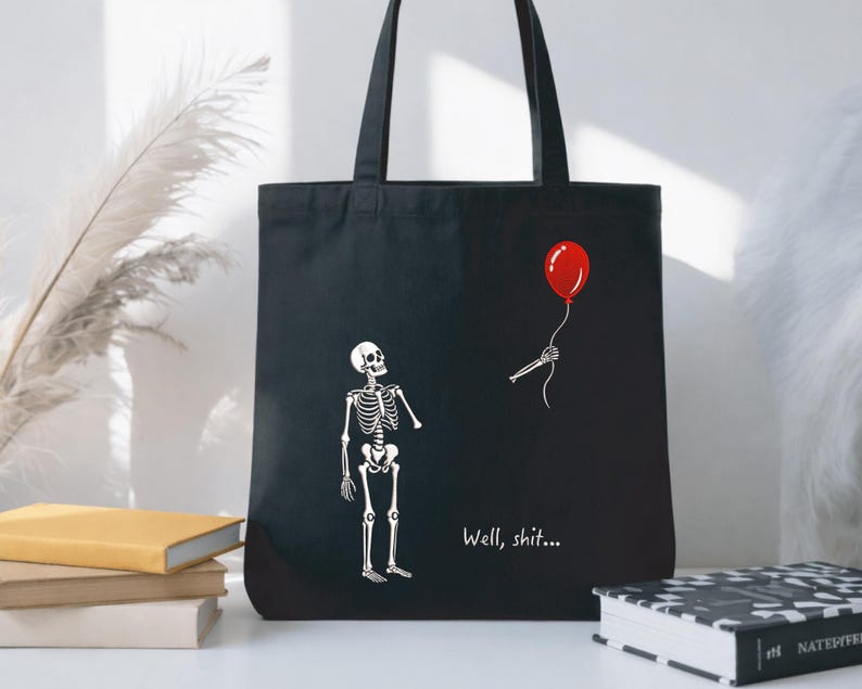 May include: A black tote bag featuring a white graphic of a skeleton looking up at a red balloon held by a disembodied skeletal hand. The text "Well, shit..." is printed in white below the skeleton. The bag has two black shoulder straps.