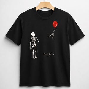 May include: A black short-sleeve t-shirt featuring a white skeleton graphic looking at a detached skeletal hand holding a red balloon. The text "Well, shit..." is printed in white below the skeleton. The shirt is displayed on a wooden hanger.