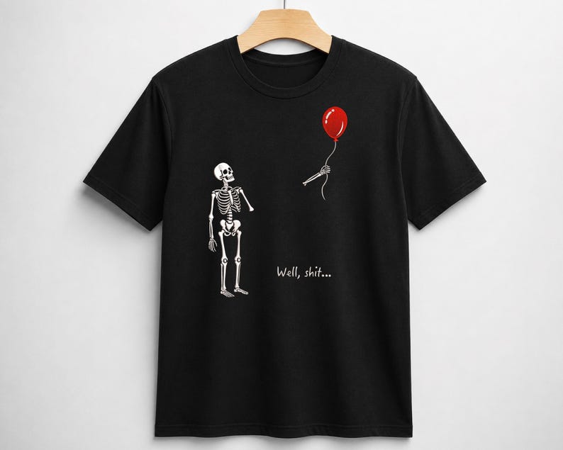 May include: A black short-sleeve t-shirt featuring a white skeleton graphic looking at a detached skeletal hand holding a red balloon. The text "Well, shit..." is printed in white below the skeleton. The shirt is displayed on a wooden hanger.