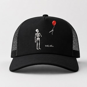 May include: A black trucker hat featuring a graphic of a white skeleton looking up at a red balloon tied to a detached skeletal hand. The text "Well, shit..." is printed in white below the skeleton on the front panel of the cap.