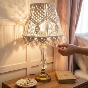 Pode incluir: Candeeiro de mesa decorativo com abajur de macramé. O abajur é creme com um padrão geométrico e franjas. A base é de metal dourado e vidro transparente.