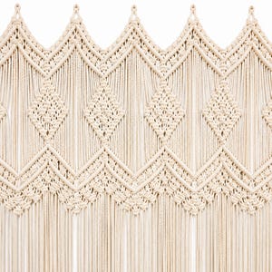 Puede incluir: Un tapiz de macramé color crema con un patrón geométrico repetitivo de diamantes y zigzags. El diseño está construido con cordón de algodón anudado, con una serie de hilos de flecos verticales que se extienden desde el borde inferior.