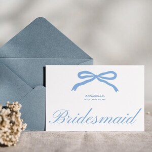 以下が含まれることがあります： 青い封筒と、"Annabelle, Will You Be My Bridesmaid"と書かれた青い文字の白いカード。カードには青いリボンのイラストが描かれています。ドライフラワーが前景にあります。