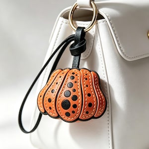 Könnte beinhalten: Ein kürbisförmiger Taschenanhänger in Orange mit schwarzen Punkten in verschiedenen Größen, befestigt an einem schwarzen Lederriemen und einem goldfarbenen Metallring, hängend an einer glatten weißen Handtasche mit goldfarbenem Druckknopf.
