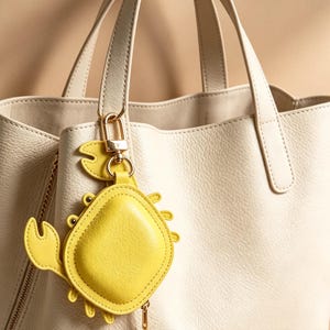 Può includere: Una borsa tote in pelle color crema con superficie testurizzata e due manici, caratterizzata da un ciondolo a forma di granchio giallo brillante attaccato a un manico con un moschettone in metallo dorato e una piccola catenella.