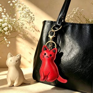 Può includere: Un portachiavi in pelle rosso brillante a forma di gatto con dettagli cuciti neri e una chiusura in metallo dorato, appeso al manico di una borsa nera testurizzata, accompagnato da una piccola statuetta di gatto in ceramica bianco sporco.