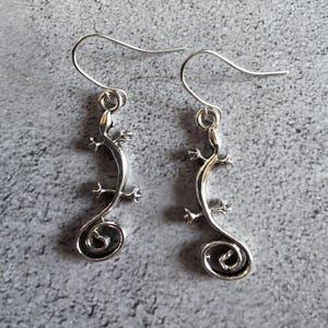May include: A pair of silver-toned lizard earrings. Each earring features a lizard design with a curled tail. The earrings have a hook closure and are displayed against a gray background.