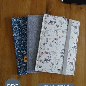 May include: Three rectangular notebook covers featuring different designs: one with a blue floral pattern and a small heart tag, one plain grey felt, and one white cover with a printed pattern of chickens, birds, and wildflowers with a grey elastic band.
