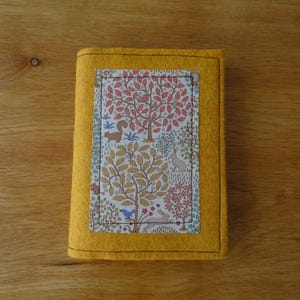 May include: A yellow felt book cover with a rectangular fabric panel featuring a tree and animal design. The fabric panel is stitched onto the cover with a black thread. The background is a wooden surface.